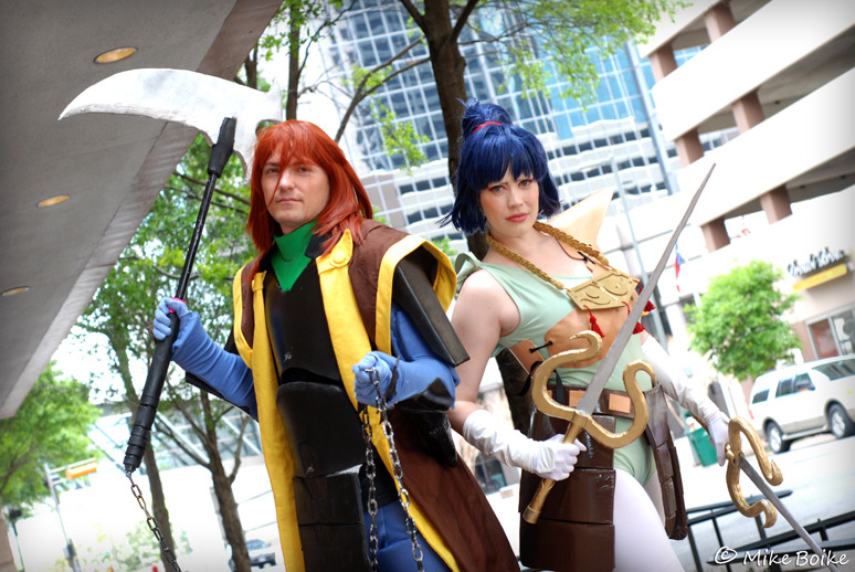 Anubis/Shuten and Kayura cosplay. Both characters in armor. Anubis/Shuten is not wearing a helmet. Characters are standing side by side, angled towards the camera.